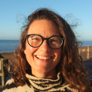 Anita is standing in front of a blurry British seaside promenade background, the sun on her face, smiling widely. She has brown curly hair, black round glasses and is wearing a warm jumper with a tall neck. She looks really happy (as she is about to go for a swim).